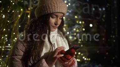美丽迷人的女孩站在一家装饰有花环的橱窗附近，使用智能手机。 新年圣诞节